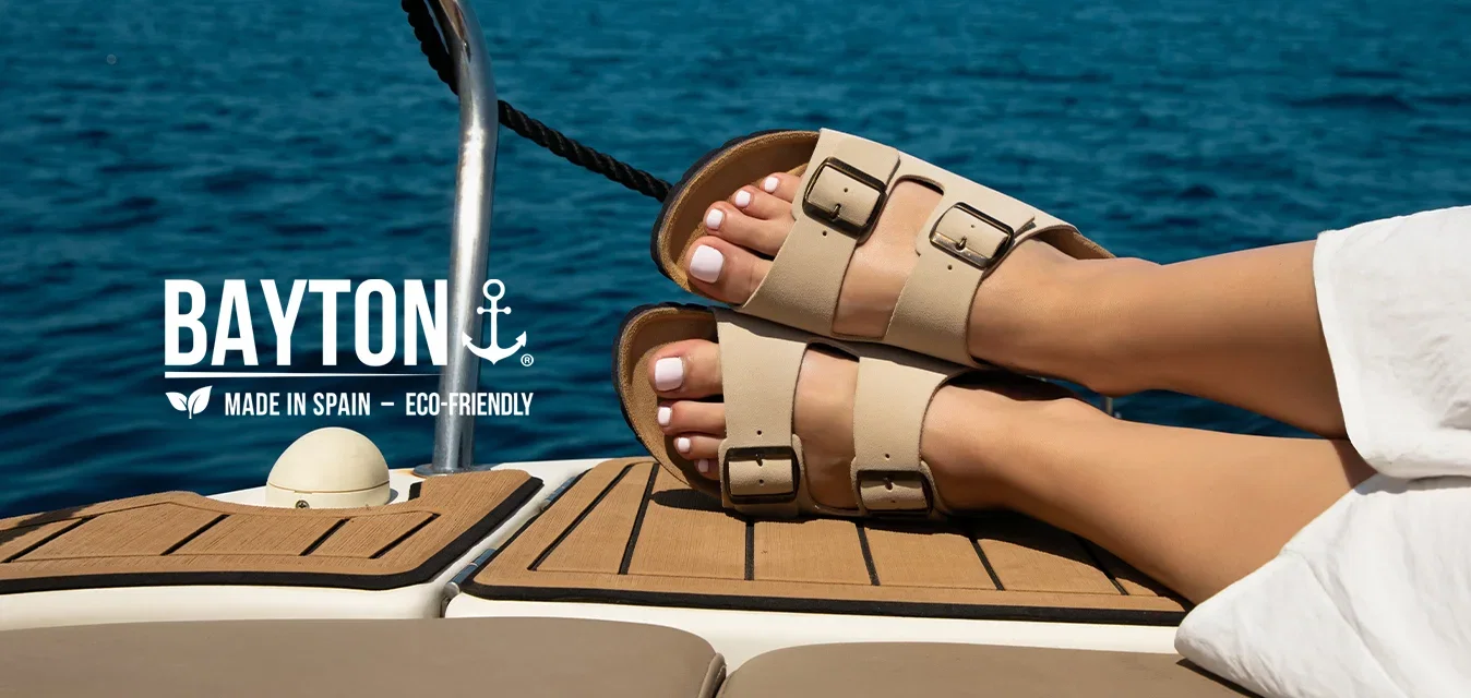 Persona que lleva sandalias beige y pantalones blancos relajándose con los pies en alto en la cubierta de un barco cerca del mar azul y tranquilo bajo un cielo despejado.