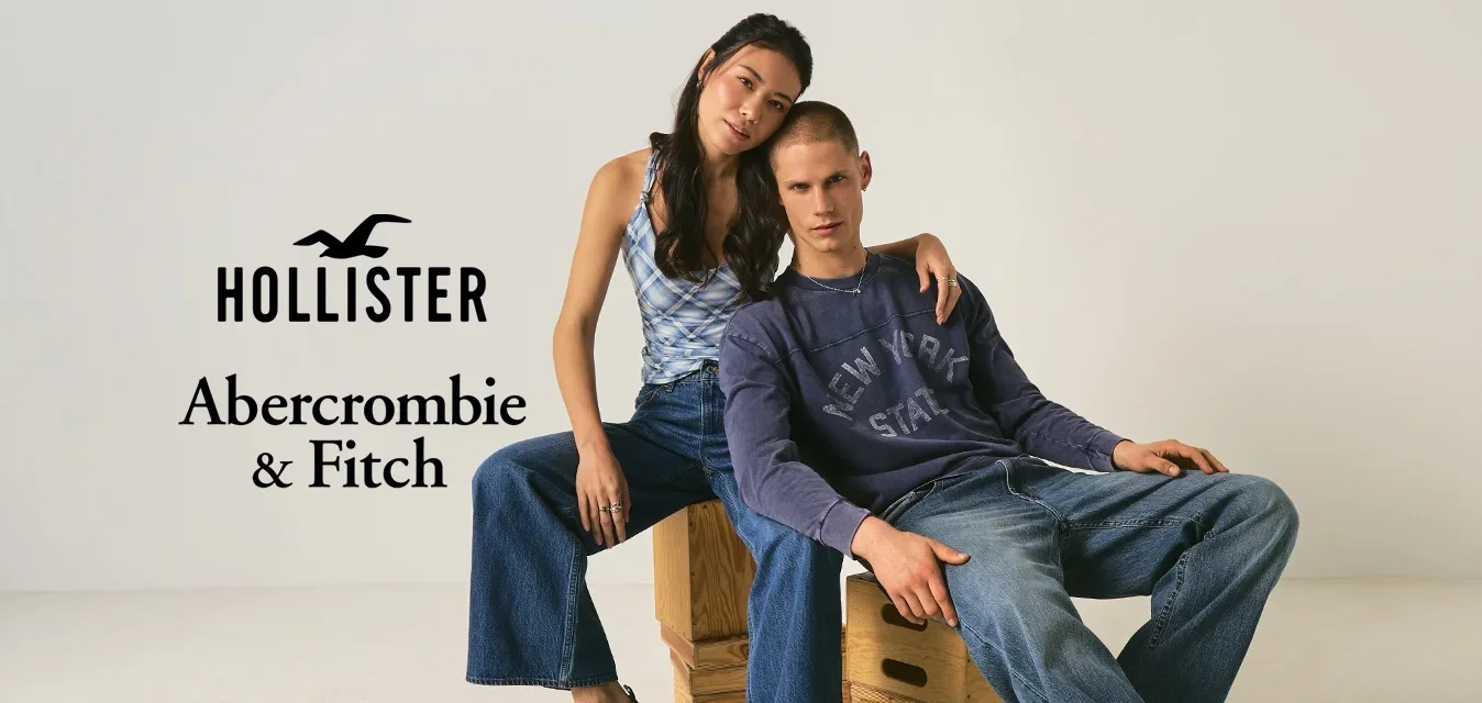 Jonge vrouw in een mouwloze top en wijde jeans zit op een houten doos met haar arm om een jonge man in een shirt met lange mouwen en jeans die naast haar zit.