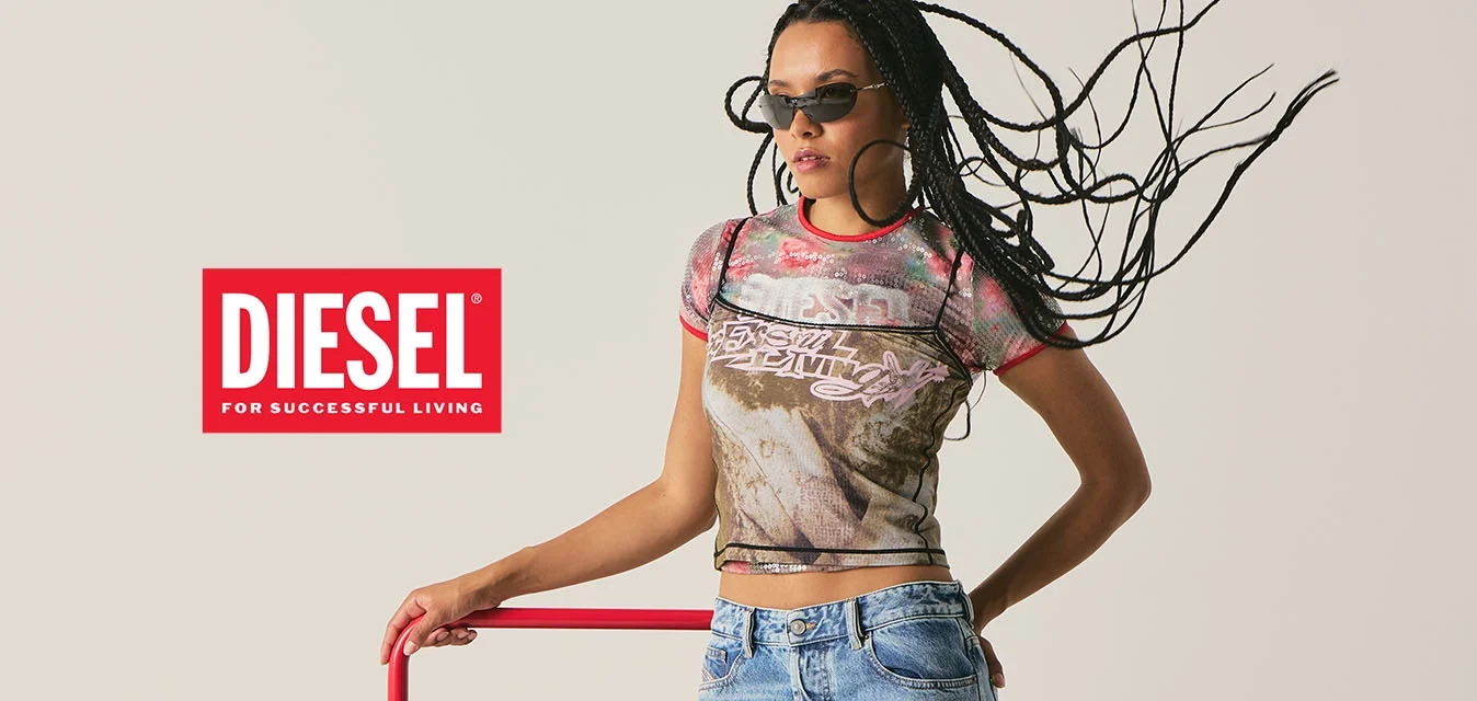 Vrouw met gevlochten haar en zonnebril, gekleed in een grafisch T-shirt en jeans, leunend op een rood metalen frame tegen een effen achtergrond.