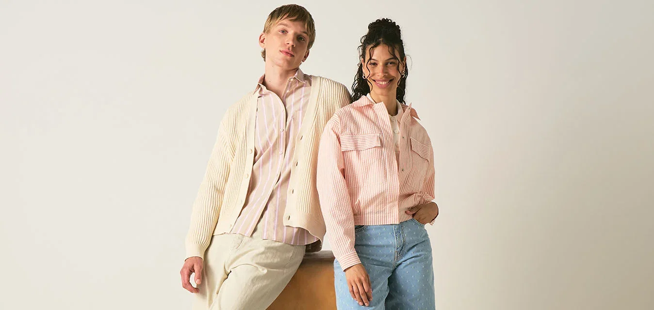 Twee jonge volwassenen in casual pastel kleding poseren, een leunt op een houten vaulting box, tegen een effen lichte achtergrond.