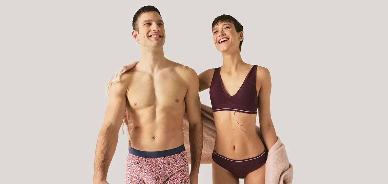 Un homme et une femme souriants en sous-vêtements se tiennent près l'un de l'autre, la femme porte une écharpe rose pâle sur le bras, arrière-plan neutre clair.
