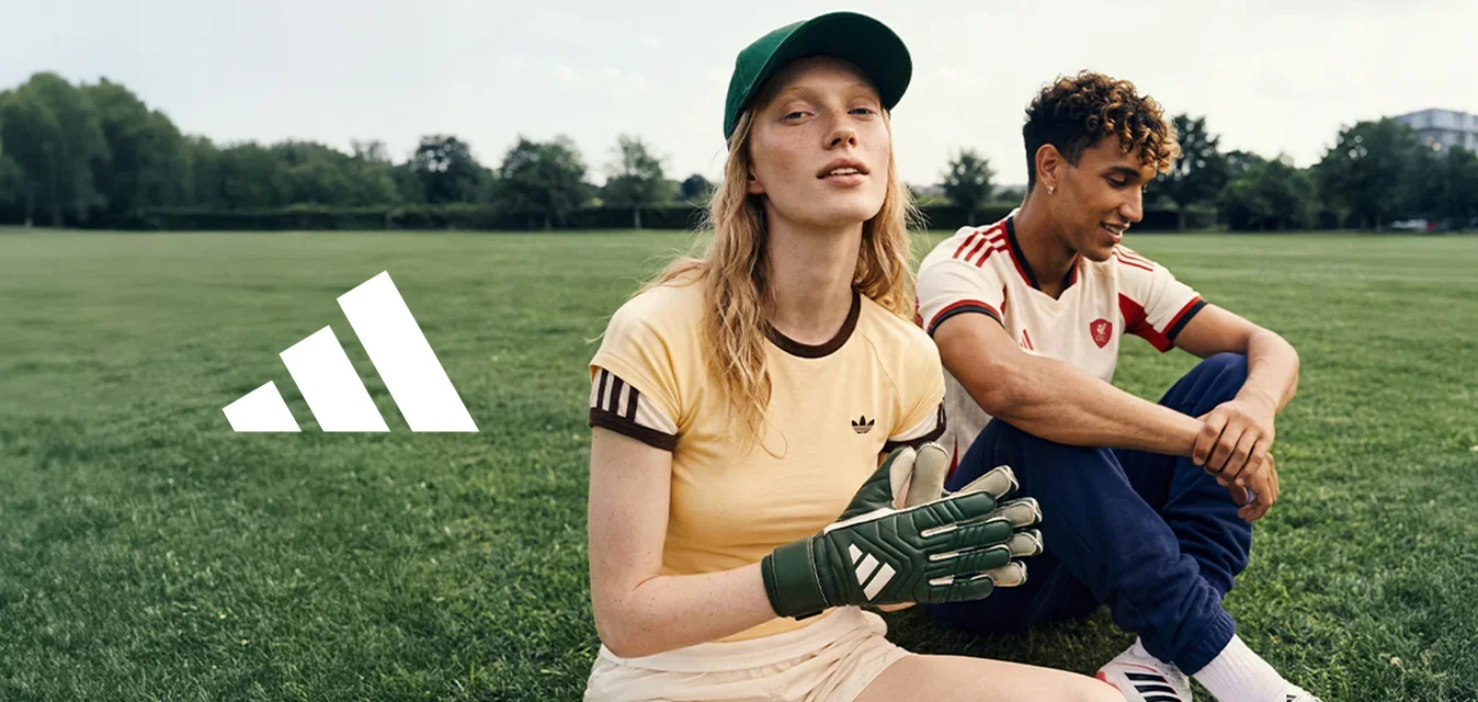 Due giovani in abbigliamento sportivo seduti sull’erba, uno indossa guanti da portiere e un cappellino, l’altro una maglia da calcio, all’aperto.