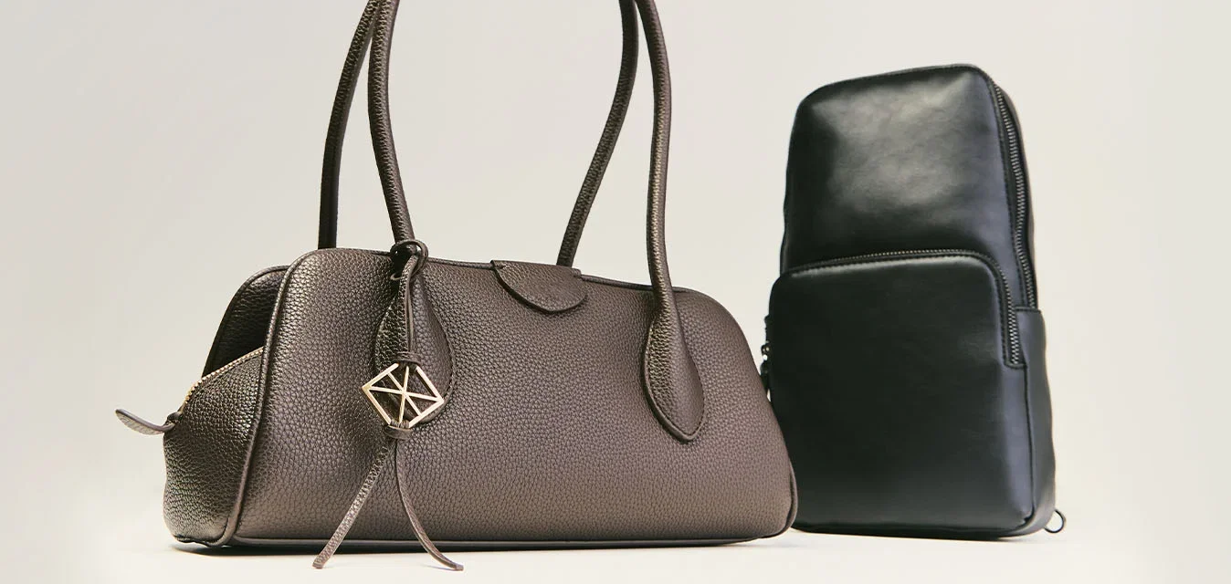 Borsa a mano in pelle marrone con doppi manici e charm geometrico accanto a una piccola borsa a tracolla in pelle nera su sfondo semplice.