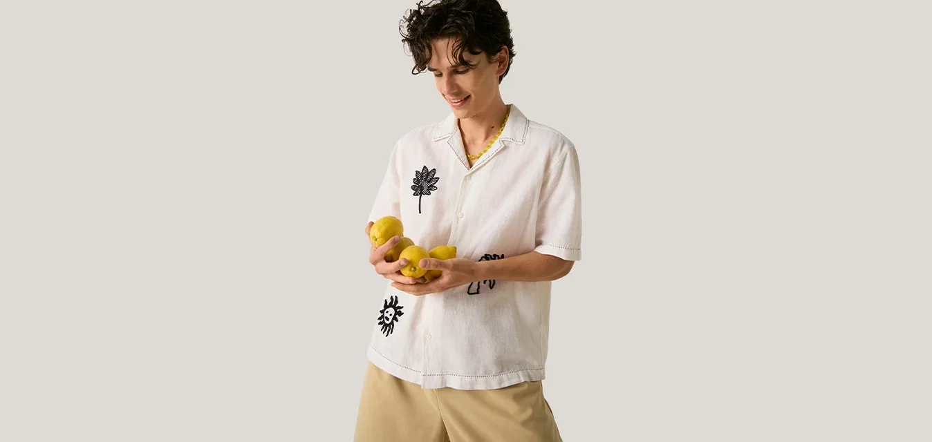 Persona con capelli corti e ricci che indossa una camicia bianca e pantaloni beige, tiene in mano diversi limoni gialli sorridendo guardandoli.