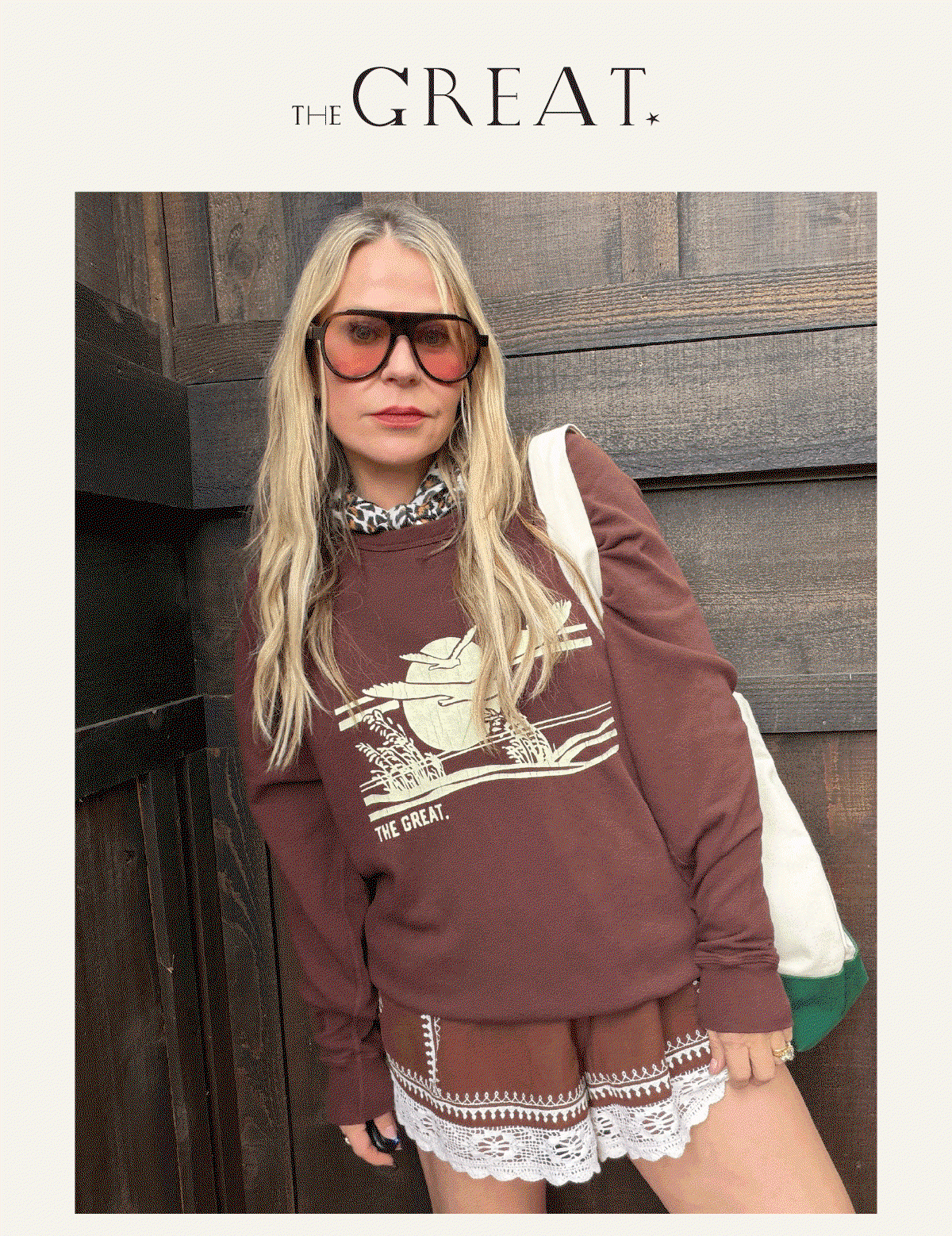 A woman wearing sunglasses, a maroon sweatshirt with a graphic design, a patterned shirt collar, and lace-trimmed shorts stands against a wooden wall with a tote bag. "THE GREAT" is written at the top of the image.