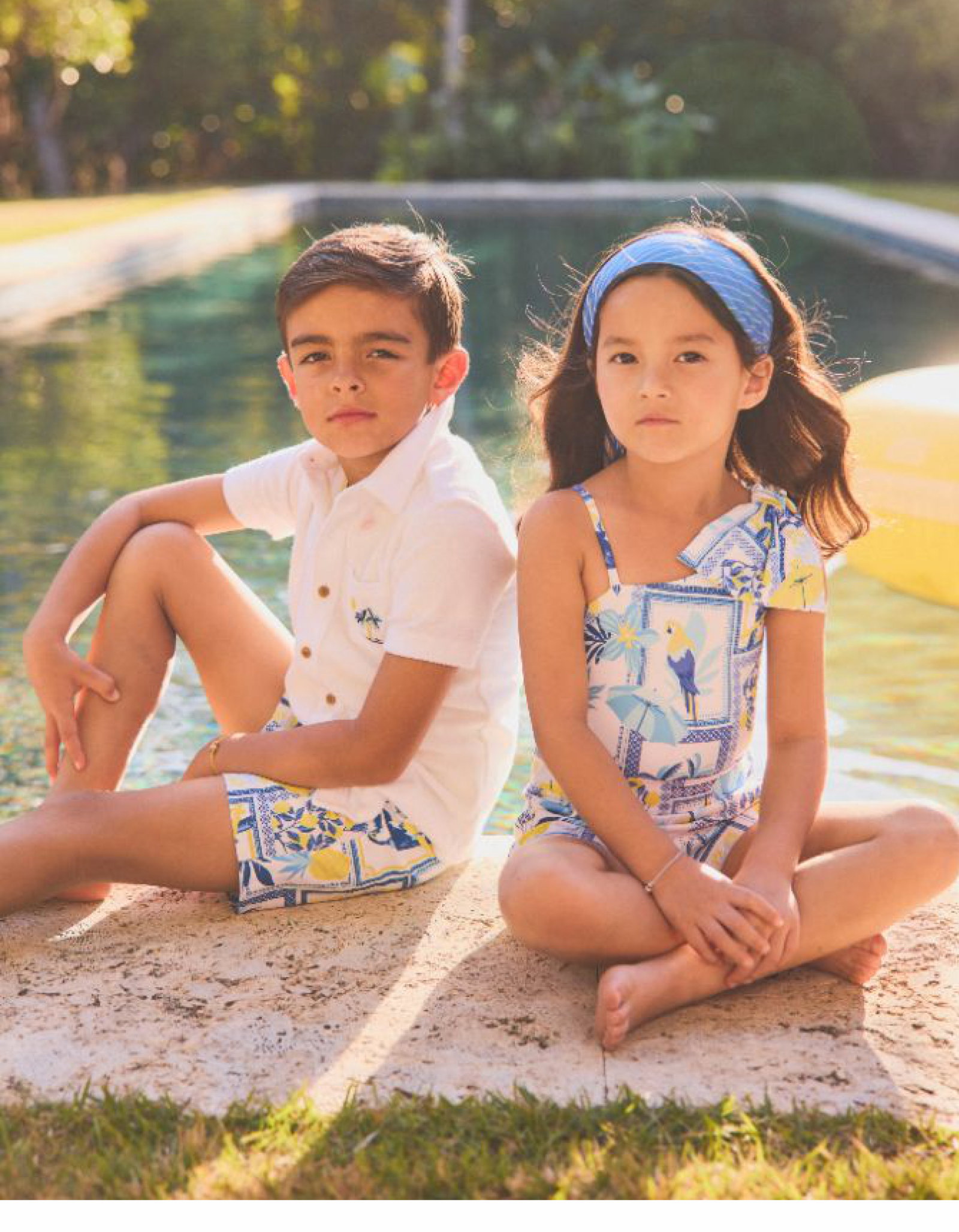 Two children sitting by a pool wearing coordinating blue, white, and yellow printed resort wear