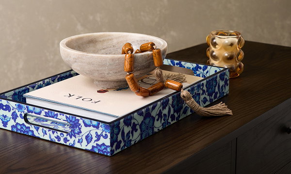 A floral display tray, a book, a candle and a bowl with a necklace inside it all sit atop a sideboard table.