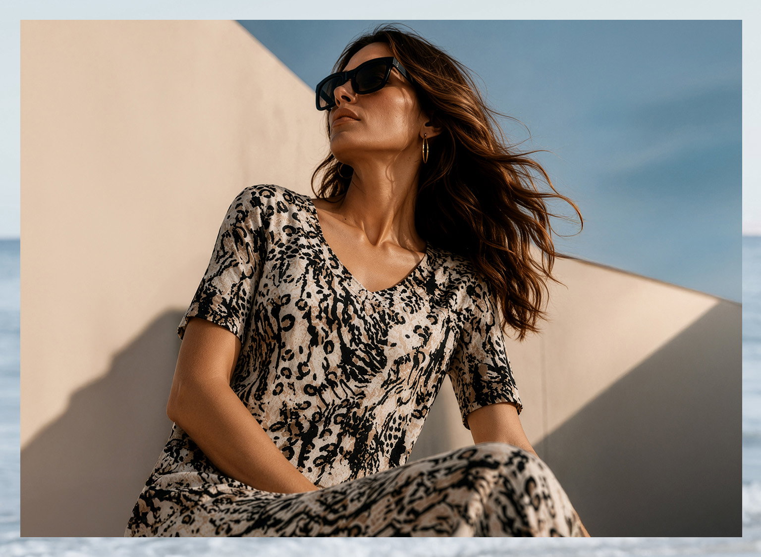 Ein Model sitzt draussen, trägt ein kurzärmeliges Kleid mit Leopardenmuster und Sonnenbrille. Der Himmel ist blau, im Hintergrund eine helle Wand. 