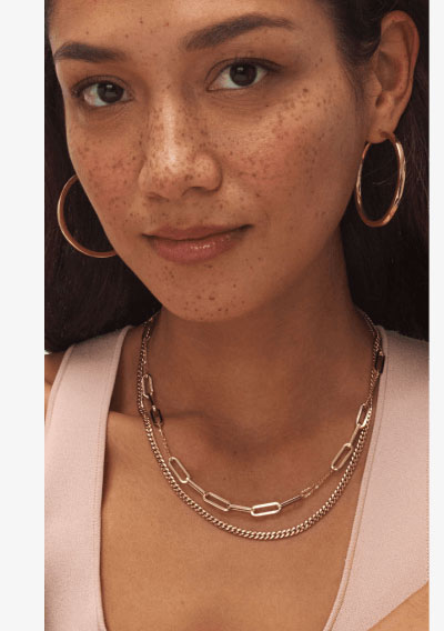 Female model wearing layered yellow gold chain necklaces and hoop earrings.
