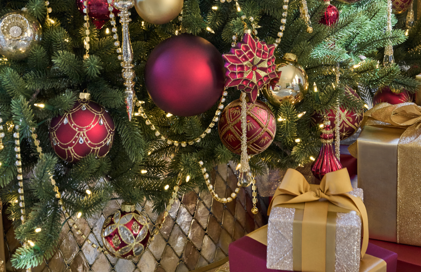 Close-up of a Christmas tree decorated with red, gold, and burgundy ornaments, beaded garlands, and wrapped gifts.