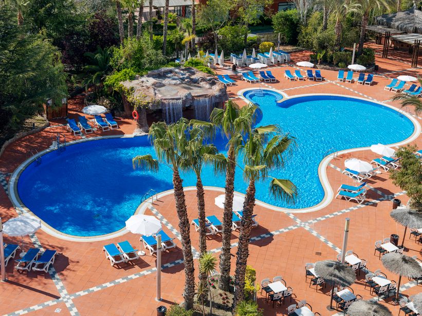 Aerial view of resort twin blue swimming pools with waterfall, palm trees, sun loungers and terracotta pool deck with parasols