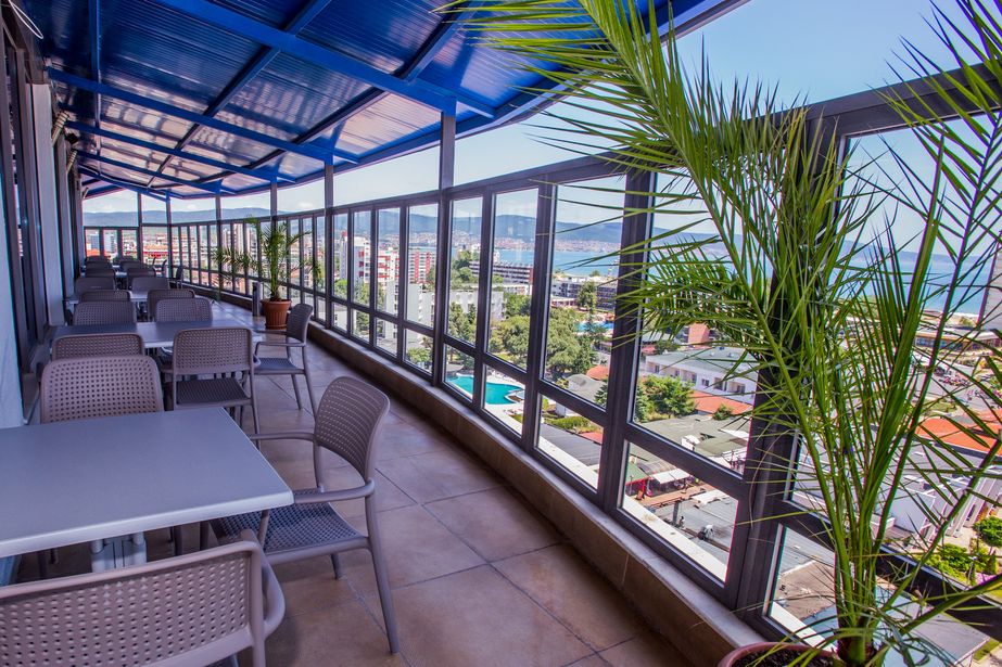 Covered rooftop terrace with tables and chairs, potted palms and glass balustrade overlooking a coastal cityscape and sea view