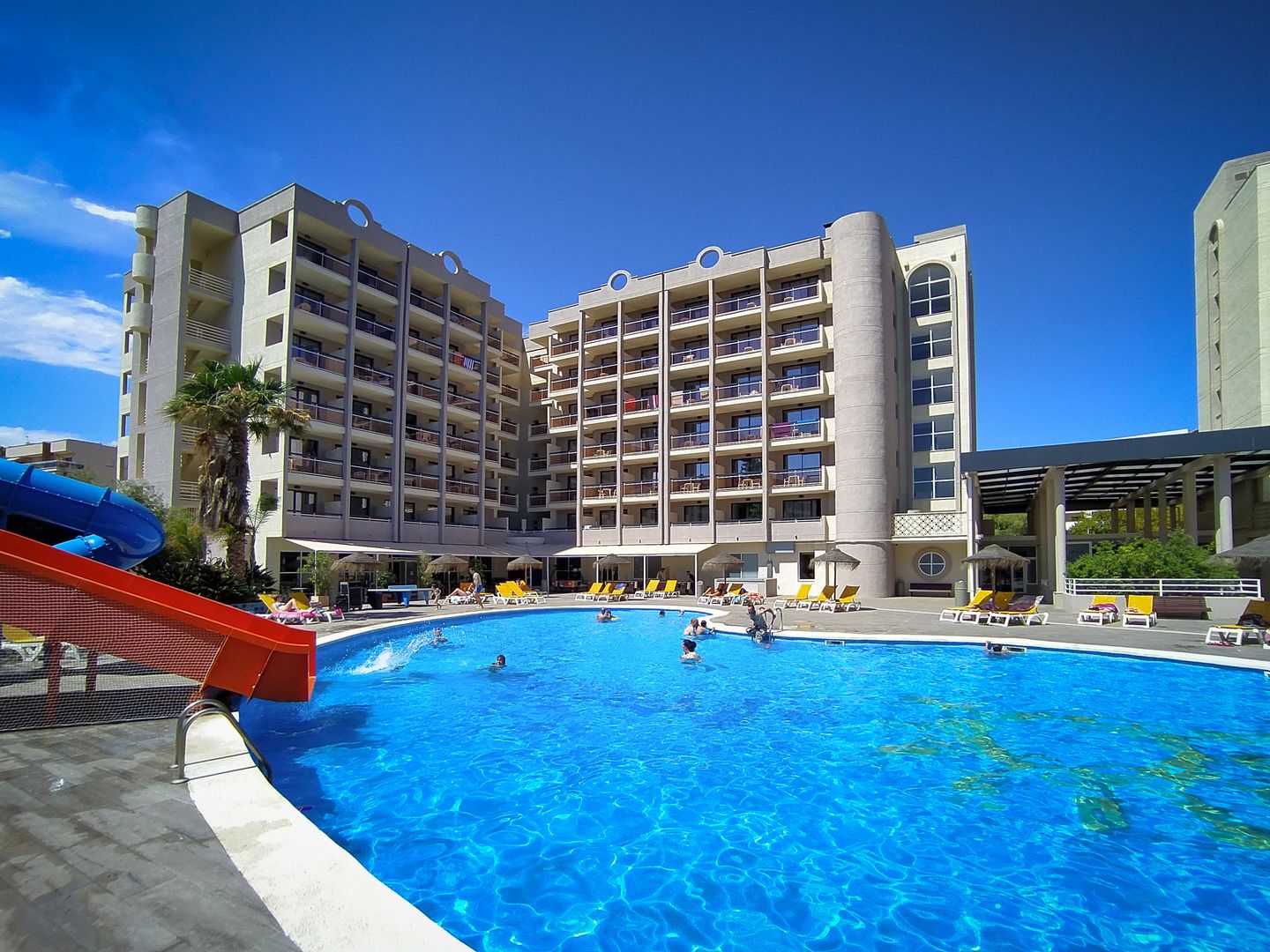 Resort hotel with large outdoor swimming pool, blue water slide, sun loungers and guests under a clear blue sky