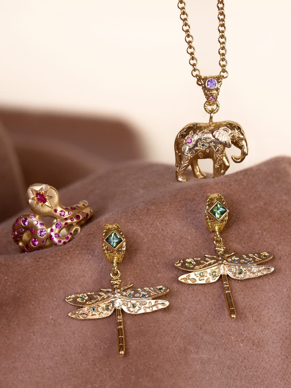 Alex Monroe limited edition Menagerie pieces. Ruby Set Snake Ring, Floral Engraved Elephant Necklace with Rainbow Sapphires and Diamonds and Floral Engraved Dragonfly Drop Huggie Hoop Earrings with Tourmalines and Blue & Green Diamonds set on a brown neutral background.