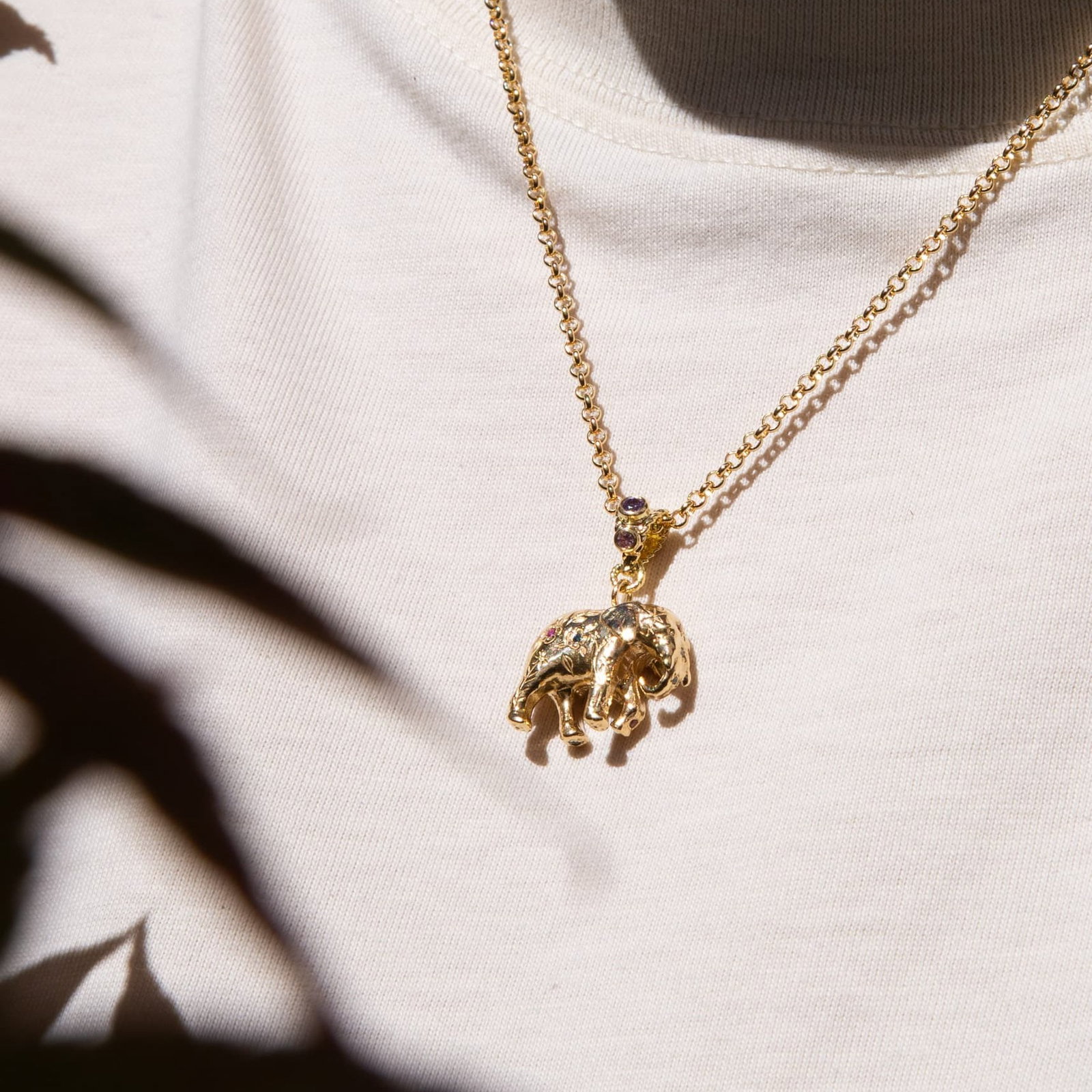 Alex Monroe Limited Edition Gold Floral Engraved Elephant Necklace with Rainbow Sapphires and Diamonds worn on a gold chain against a white top, in natural sunlight.