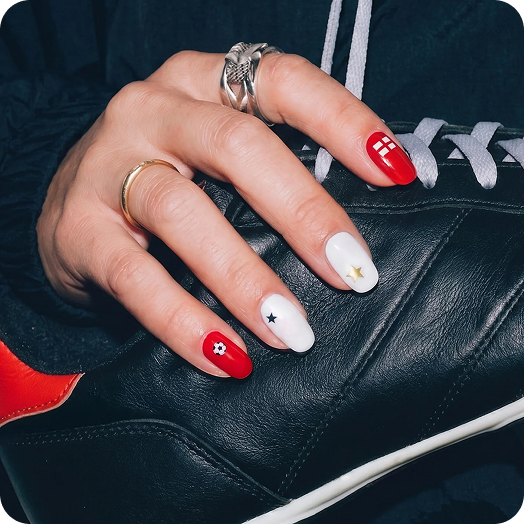 Close-up of a hand with red and white manicured nails featuring a soccer ball design, a black star, and a small flower accent, resting on a black leather bag with white laces