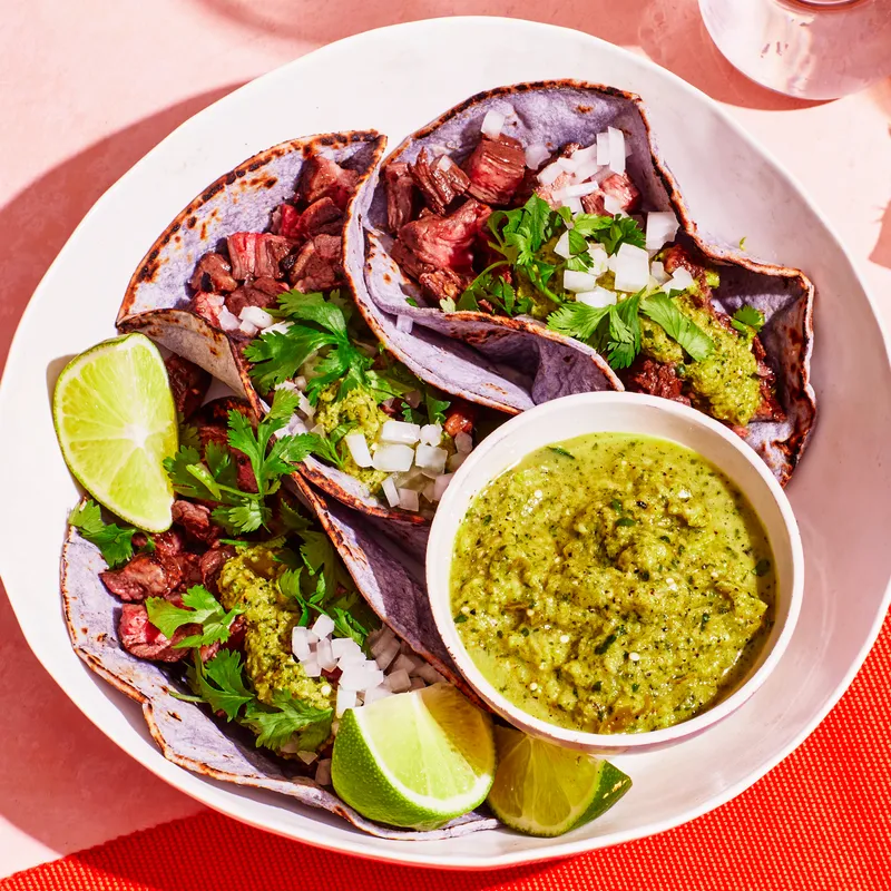 Plate with three carne asada tacos and a bowl of salsa verde over a pink background