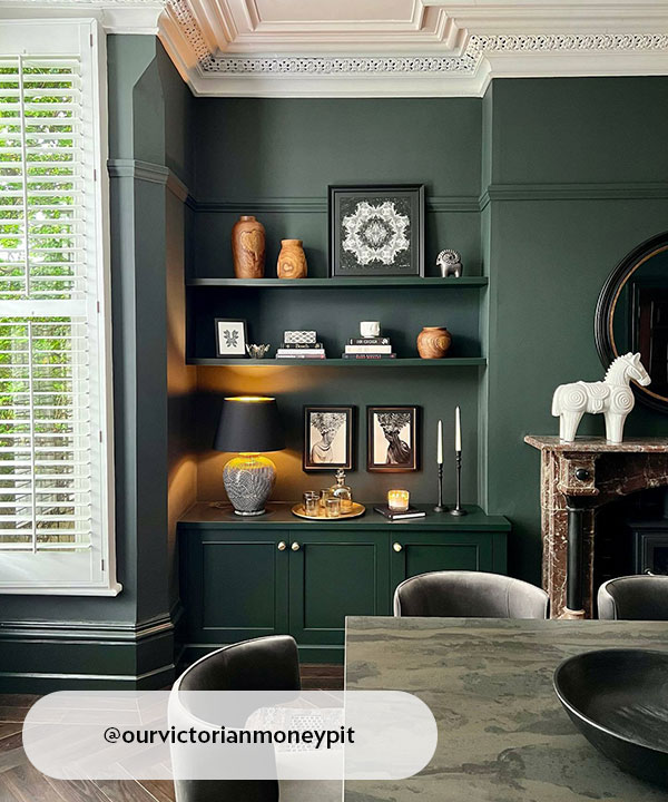 A dramatic dining area with deep forest green walls and a grand marble fireplace. Built-in alcove shelving is painted to match the walls, displaying curated ornaments under warm atmospheric lighting.