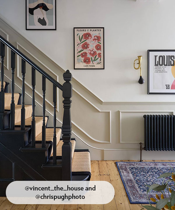 A classic staircase with a black-painted banister and a natural sisal runner. The walls feature off-white decorative picture frame moulding, complemented by a black period-style radiator and polished wood flooring.