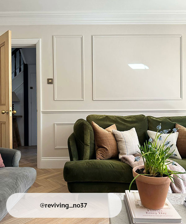A bright living room with elegant cream walls and decorative wall moulding. An olive green velvet sofa is styled with neutral cushions, set against a backdrop of light oak herringbone flooring and a matching wooden door.