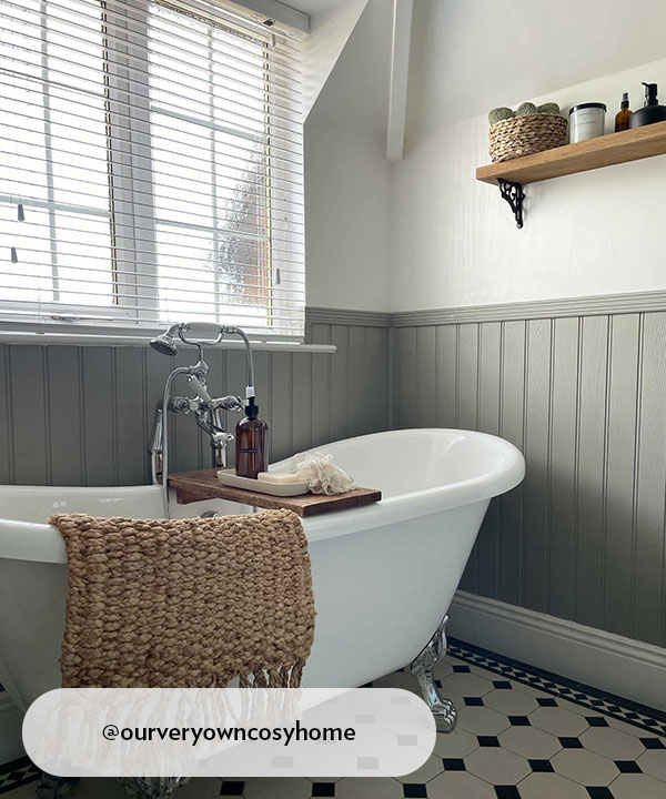 A traditional roll-top bathtub with chrome claw feet and a vintage-style mixer tap. The lower half of the wall features sage green tongue-and-groove panelling, with a wooden bath caddy and a woven rug adding natural textures.
