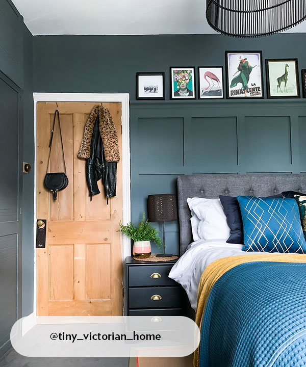 A sophisticated bedroom featuring a dark teal panelled feature wall and a grey button-back headboard. Above the bed, a narrow picture ledge displays various framed art prints, and a natural wood door adds warmth to the space.