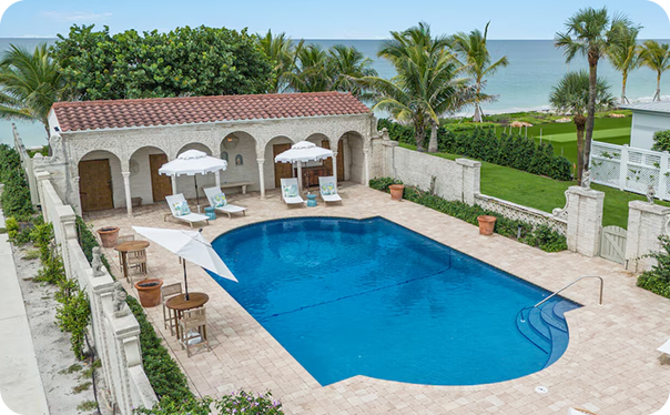 Courtyard pool with lounge chairs and umbrellas beside an arched cabana, palm trees and ocean beyond.