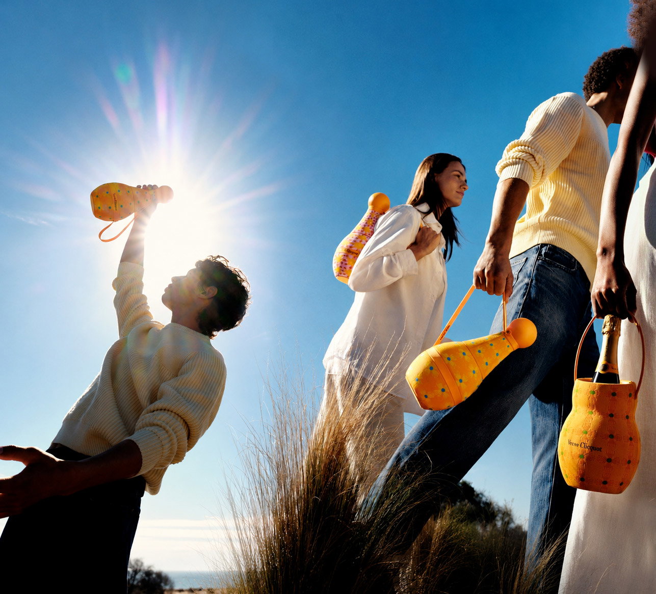 Group of friends walking on a sunny dune carrying yellow dotted champagne coolers and bottles under a blue sky