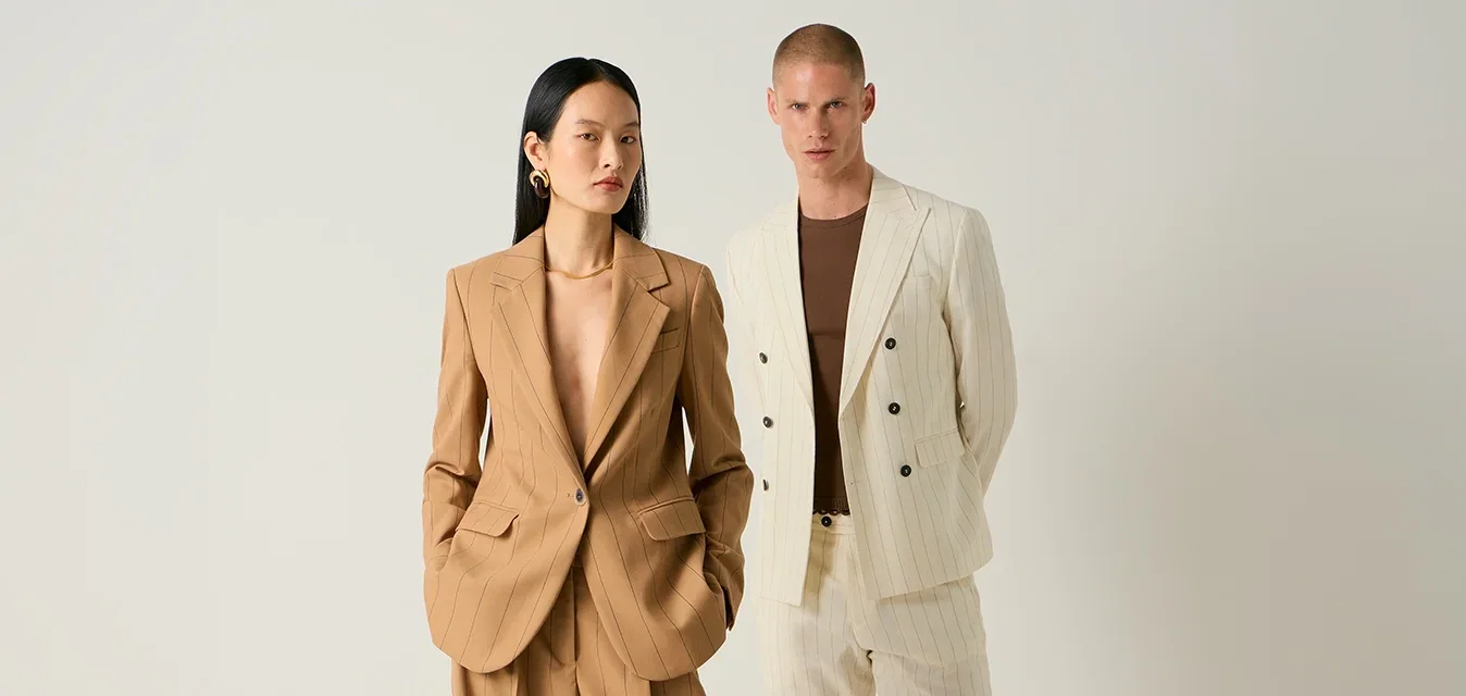 Due modelli sono in posa su uno sfondo semplice, uno con un tailleur pantalone a righe beige, l’altro con un completo crema a righe, camicia marrone e scarpe nere.