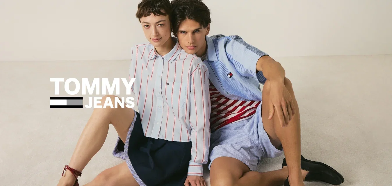 Due giovani persone sono sedute sul pavimento appoggiate l'una all'altra, indossando camicie a righe casual, pantaloncini e mocassini.
