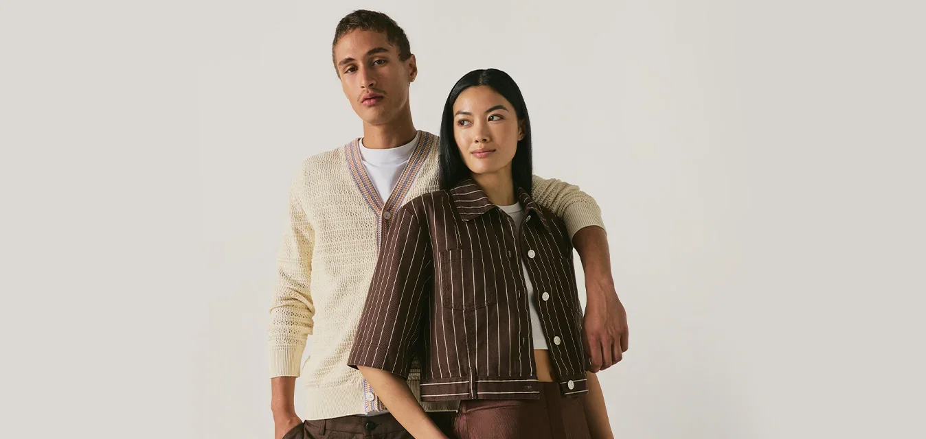 Due persone stanno insieme; una indossa un cardigan crema e pantaloni marroni, l'altra una giacca a righe marrone e una gonna. Entrambe guardano avanti con sicurezza.