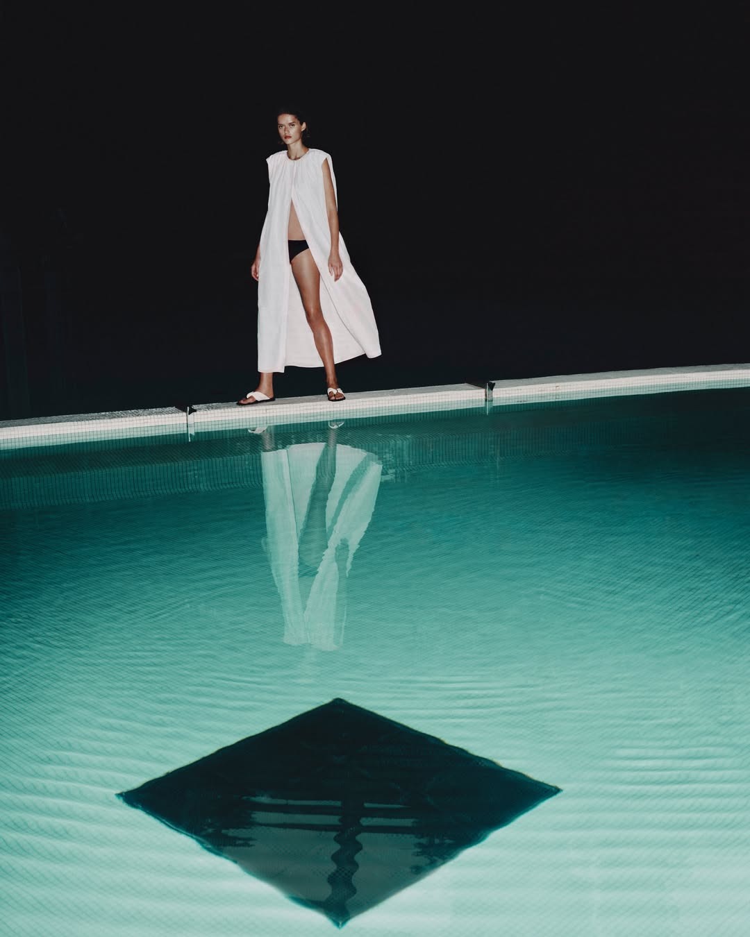 Woman in white flowing cover-up and black swimwear standing by an illuminated pool at night, reflected in the still water