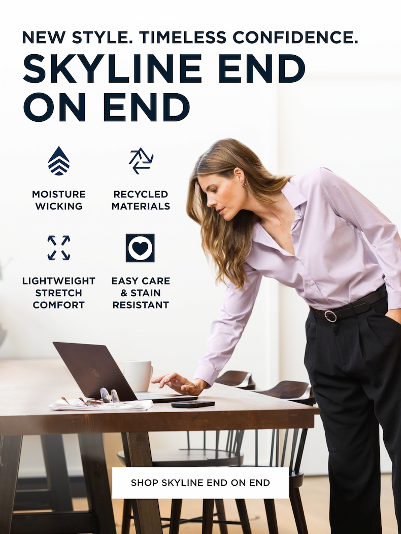 Woman wearing Skyline button down shirt and black trousers standing up at the dining table and looking at her laptop with coffee and glasses beside her. Shop Skyline End on End.