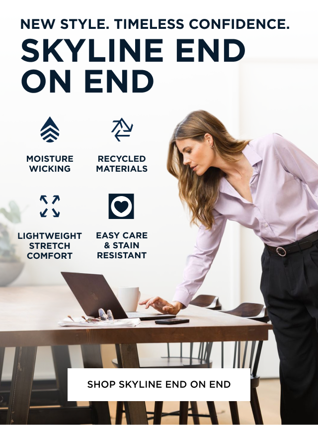 Woman wearing Skyline button down shirt and black trousers standing up at the dining table and looking at her laptop with coffee and glasses beside her. Shop Skyline End on End.