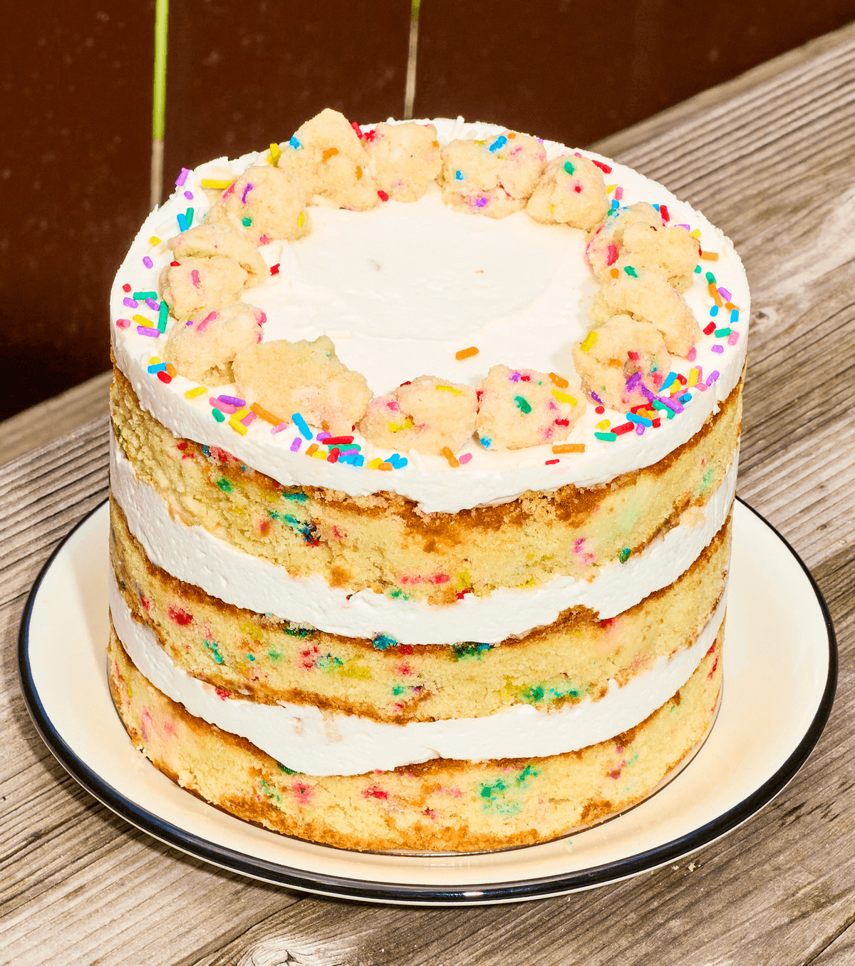 Three-layer confetti birthday cake with white frosting, rainbow sprinkles, and a ring of cake crumbs on top, displayed on a white plate.