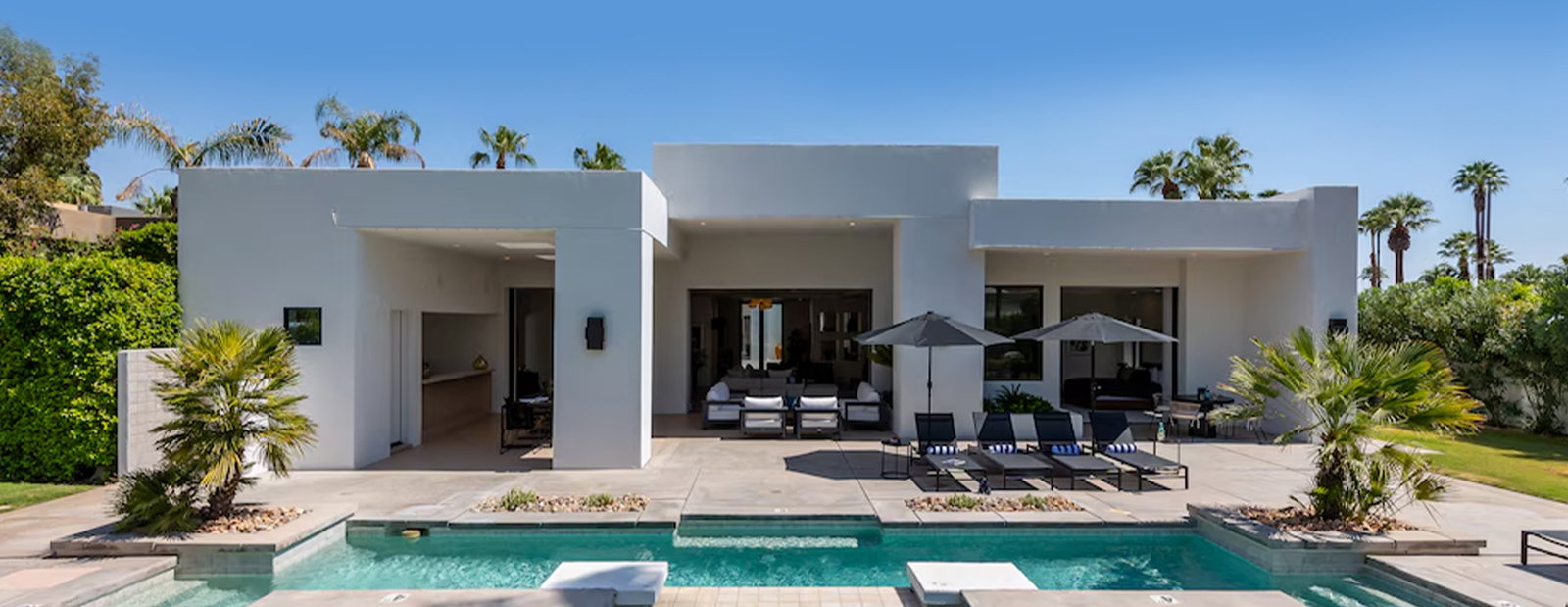Modern vacation home with a pool, lounge chairs, umbrellas, and palm trees in a sunny outdoor setting.