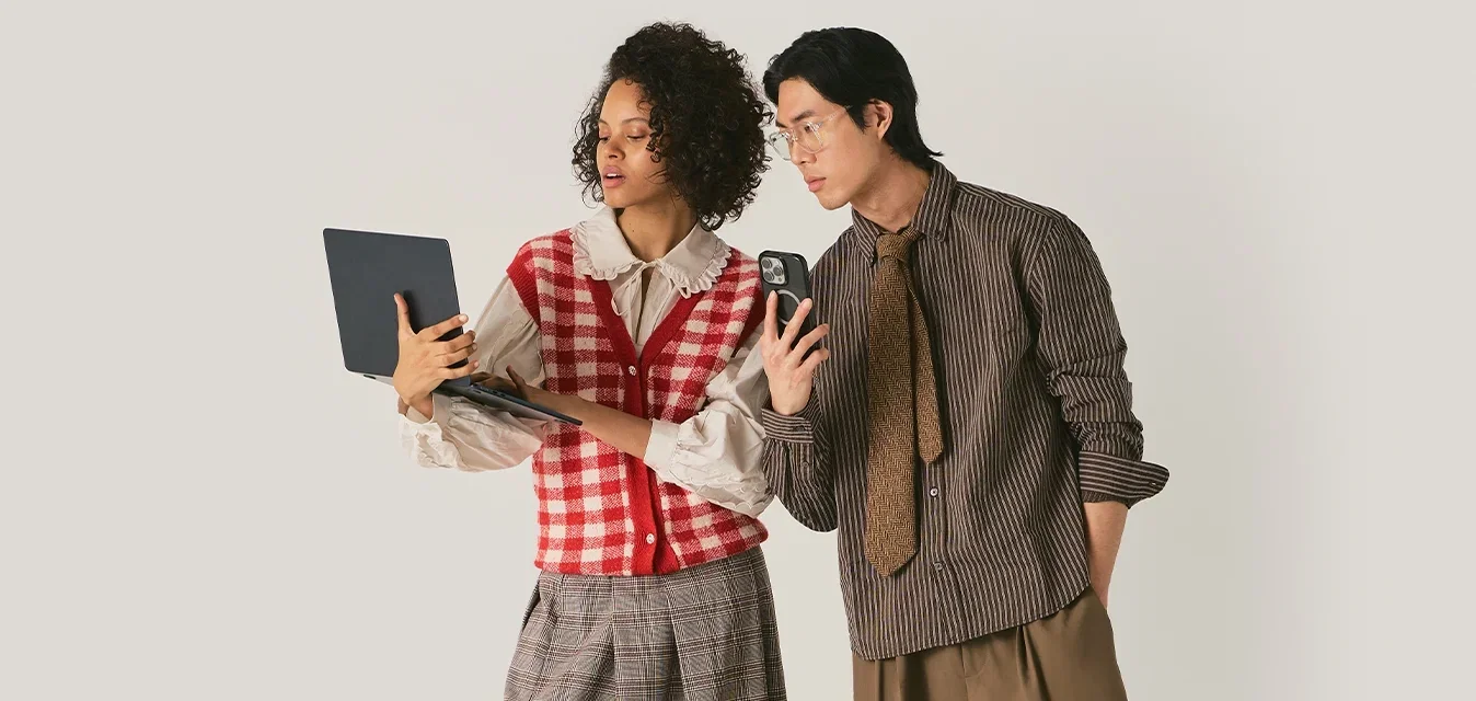 Dos personas de pie mirando una laptop y un teléfono, vestidas con ropa de patrones sobre un fondo liso.