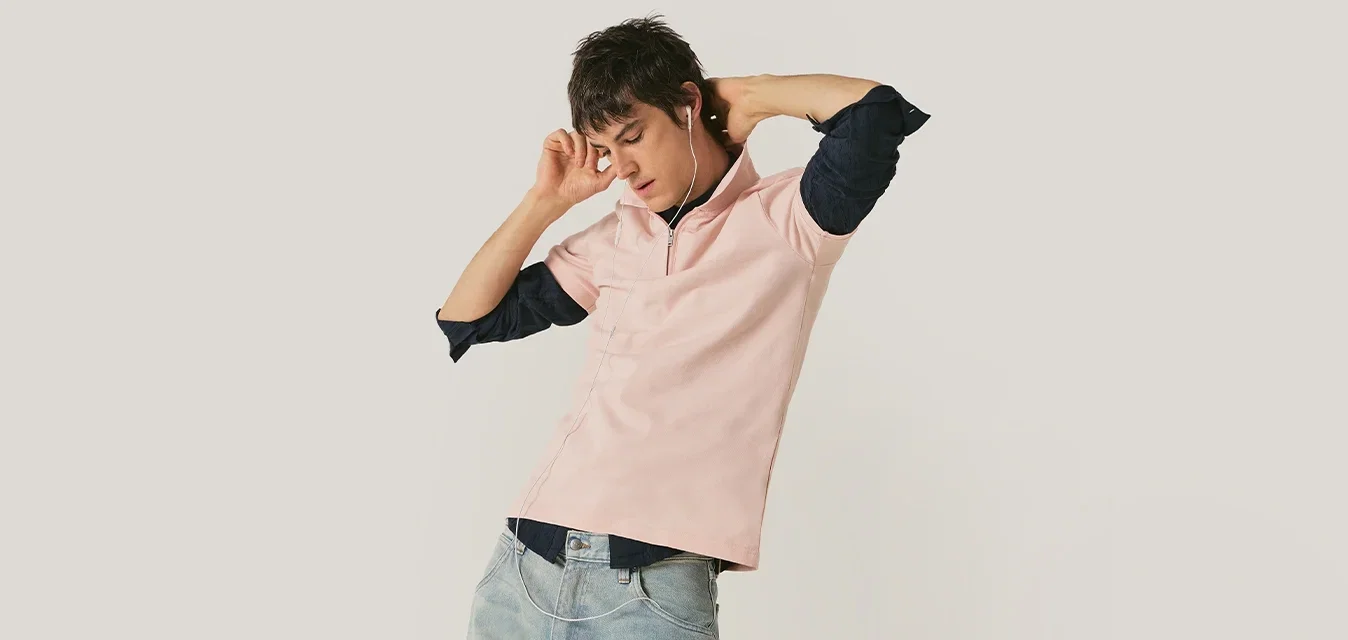 Joven con camisa rosa claro y vaqueros estira los brazos detrás de la cabeza contra un fondo liso y claro.