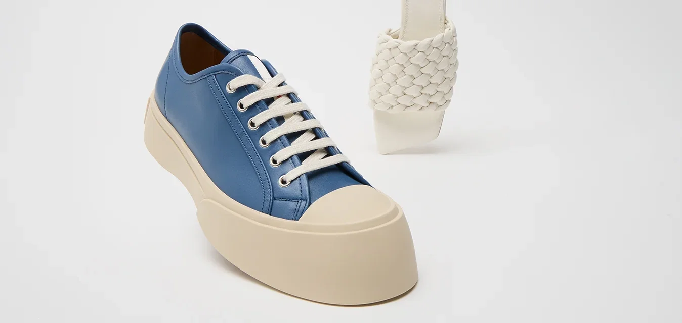 Zapatilla de cuero azul con cordones blancos y una suela gruesa beige; al lado, una sandalia deslizante blanca trenzada con un tacón curvado.