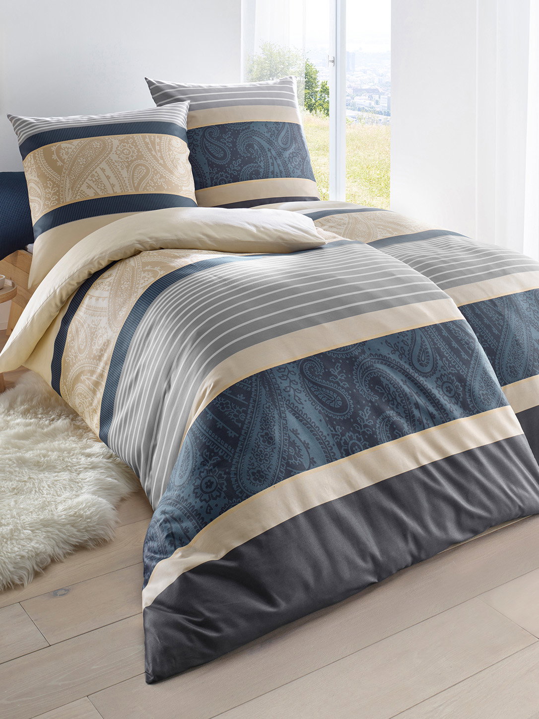  Ein Bett ist mit Bettwäsche in Streifenmuster bezogen, die Farben sind Blau, Beige, Grau und Weiss mit Paisley-Details. Das Zimmer ist hell mit einem Fenster und einem weissen Teppich.