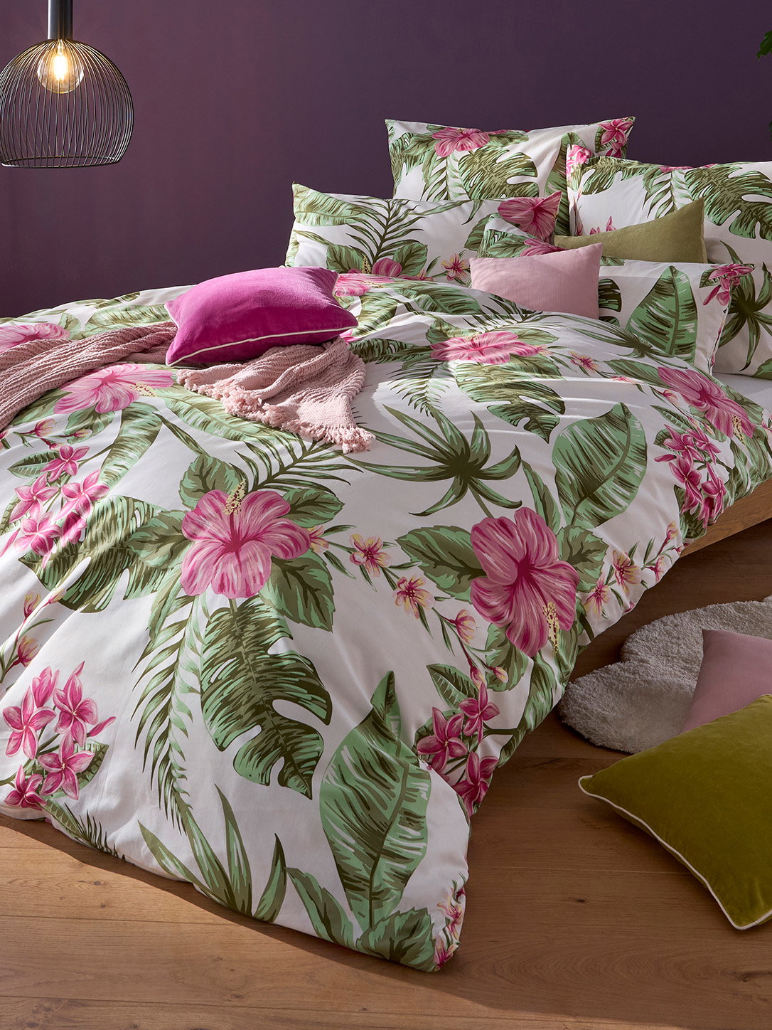 Ein Bett ist mit Bettwäsche in Weiss mit grossem grünem Blatt- und pinkem Blumenmuster bezogen. Verschiedene Kissen in Rosa und Grün liegen darauf. Im Hintergrund ist eine violette Wand.