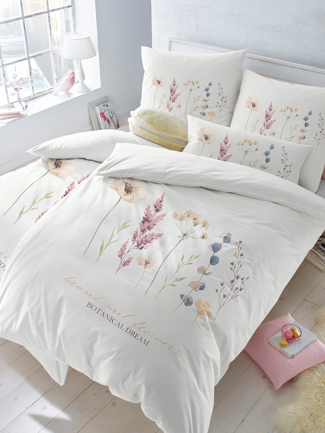 Ein Bett ist mit weisser Bettwäsche mit zarten, bunten Blumenmotiven bezogen. Die Kissen haben das gleiche florale Design. Das Zimmer ist hell und freundlich mit einem Fenster und einem kleinen Nachttisch.