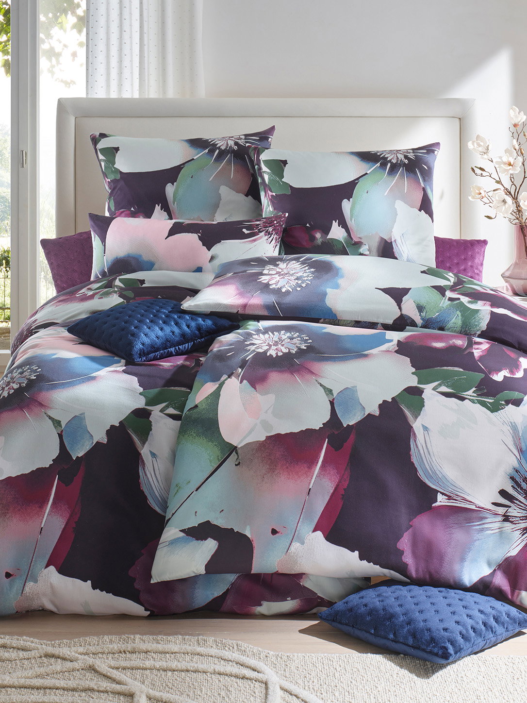 Ein Bett ist mit Bettwäsche in dunklen Farben mit großem floralem Muster bezogen. Dazu liegen dunkelblaue und violette Kissen darauf. Das Bett steht vor einem weißen Kopfteil und einem Fenster.