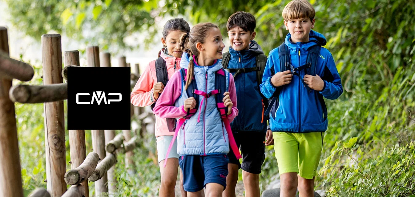 Quatre enfants avec des sacs à dos marchent sur un sentier forestier à côté d'une clôture en bois, entourés d'arbres verts et de feuillage.