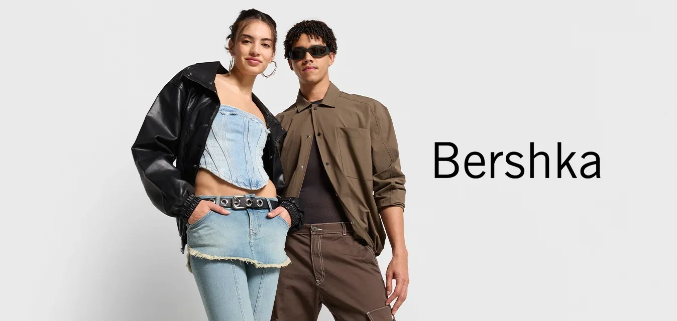 Deux individus se tiennent côte à côte. L'un porte une veste en cuir noire et un haut en jean cropped avec un pantalon taille haute. L'autre porte une chemise brune et un pantalon.
