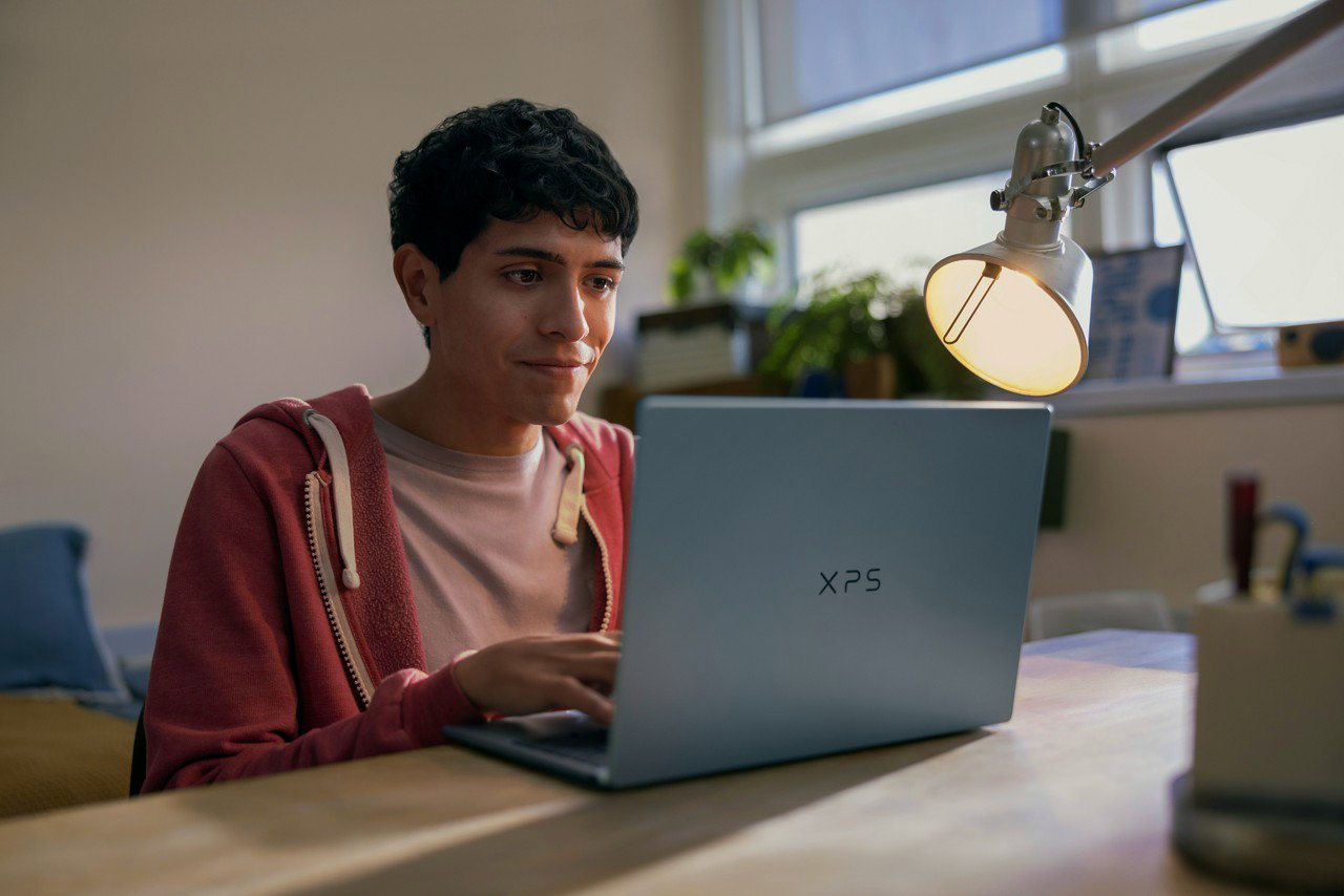 Student using a Dell XPS laptop at a desk under a lamp in a cozy study environment.