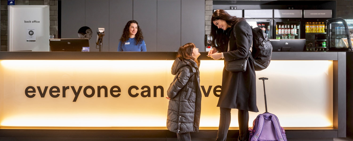 Kind spricht mit Mutter an der a&o Hostelrezeption, 'everyone can travel' Schriftzug auf dem Tresen