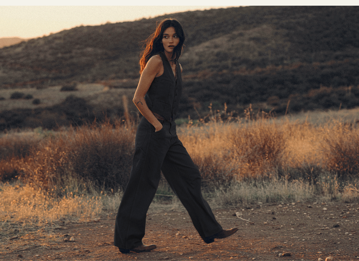 Woman wearing a washed black tailored vest and wide-leg pants, walking through a sunlit desert landscape at golden hour.