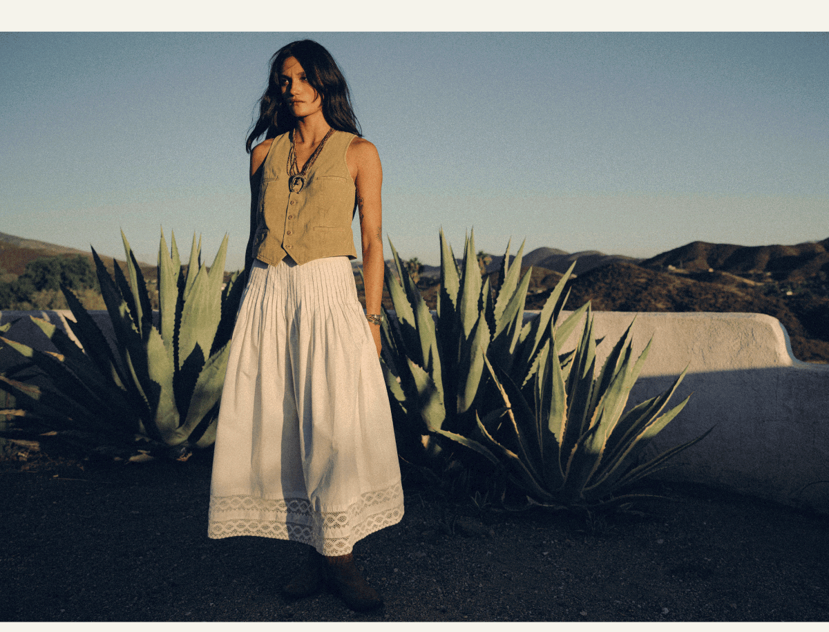 Woman wearing a tan tailored vest and flowing white lace-trimmed skirt, standing among agave plants against a sunlit desert landscape.