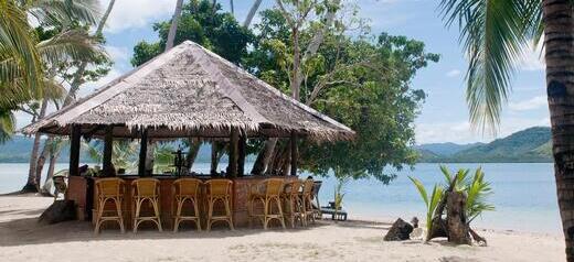 Wicker bar stools surround a beach bar with the turquoise sea in the background. 
